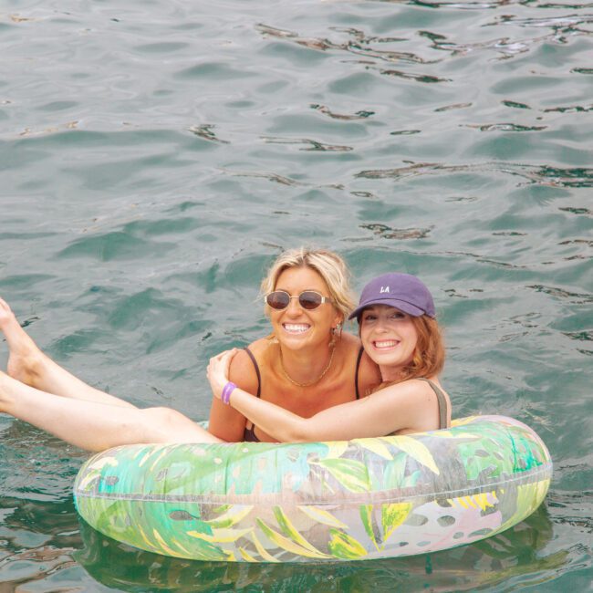Two smiling women float together in a colorful inflatable tube on clear, rippling water. One wears sunglasses and the other a cap. Both look happy and relaxed, enjoying a sunny day outdoors.