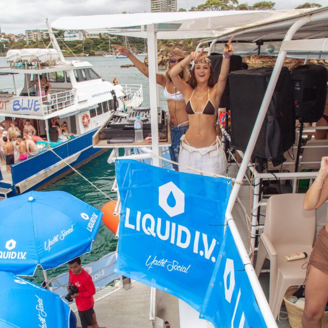 A group of people in swimwear celebrate on boats decorated with "LIQUID I.V." banners and blue umbrellas, with some dancing and others socializing near the water on a cloudy day.