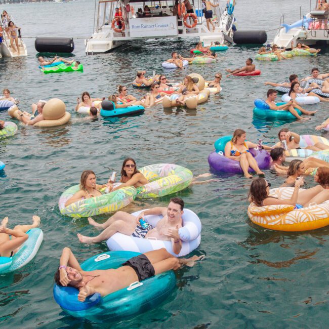 A large group of people relax on colorful inflatable floats in a lake, surrounded by boats. Many are chatting, smiling, and enjoying the sunny day on the water.