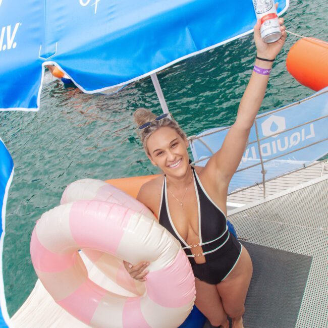 A smiling woman in a black swimsuit holds a can up and a pink-and-white swim ring under her arm, standing on a dock by the water under blue Liquid I.V. umbrellas.