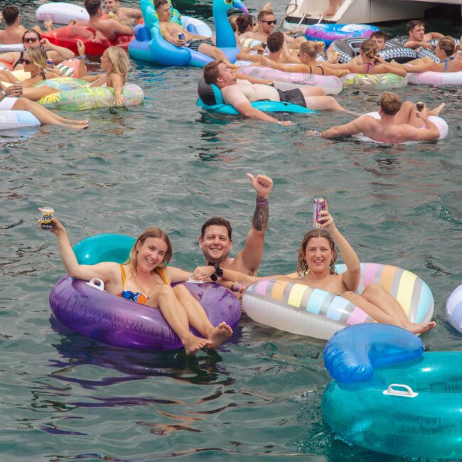 A group of people relaxing on colorful inflatable floats in the water, smiling and holding drinks, with others floating and socializing nearby; a lively summertime scene.