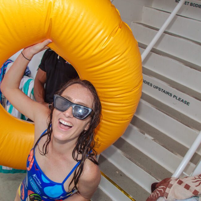 A smiling woman in sunglasses and a swimsuit holds a large yellow pool float, standing near white stairs with towels and "No Wet Bodies Upstairs Please" signs visible.