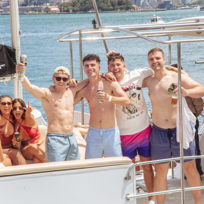 A group of young people enjoys a sunny day on a boat; four men stand shirtless at the front smiling with drinks, while others sit and laugh together. The water and shoreline are visible in the background.