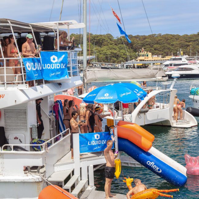 People enjoy a lively boat party with music, drinks, and pool floats on a sunny day. Some are on the boat, others swim or use a waterslide into the water. Nearby boats and a wooded shoreline are in the background.