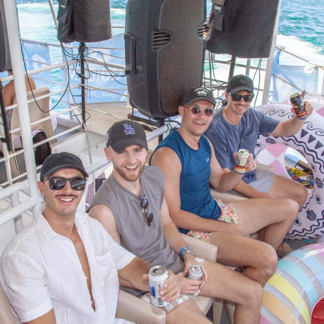 Four men sit and smile on a boat, each holding a canned drink. They wear casual summer clothing, sunglasses, and hats. Behind them are speakers, pool floats, and blue water in the background.