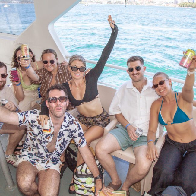 A group of seven people smile and raise cans while sitting on a boat, enjoying sunny weather with blue water and a city skyline in the background. They wear summer clothes and sunglasses, celebrating together.