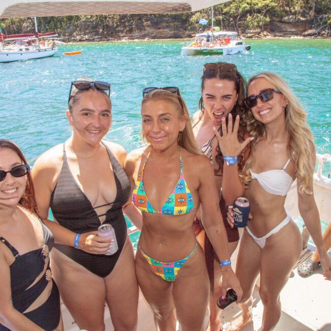 Five women in swimsuits stand and smile on a boat, holding drinks, with blue water and a forested shoreline in the background. It’s a sunny day and other boats are visible in the distance.