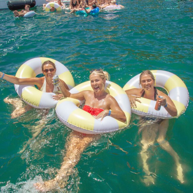 Three women in swimsuits smile and hold drinks while floating in yellow and white inflatable rings on bright blue water, with other people and floaties in the background at a lively outdoor event.
