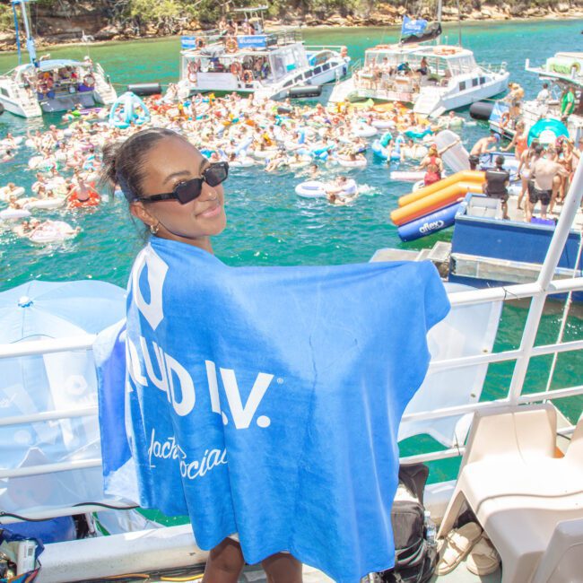 A smiling woman wearing sunglasses and a blue towel stands on a boat deck, overlooking a lively packed floating party with people on inflatables and surrounding boats in a scenic bay.