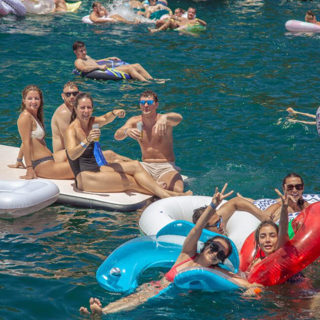 A group of young adults relax and pose on colorful pool floats and an inflatable dock in a vibrant blue lake, smiling, making peace signs, and enjoying a sunny day surrounded by others in the water.
