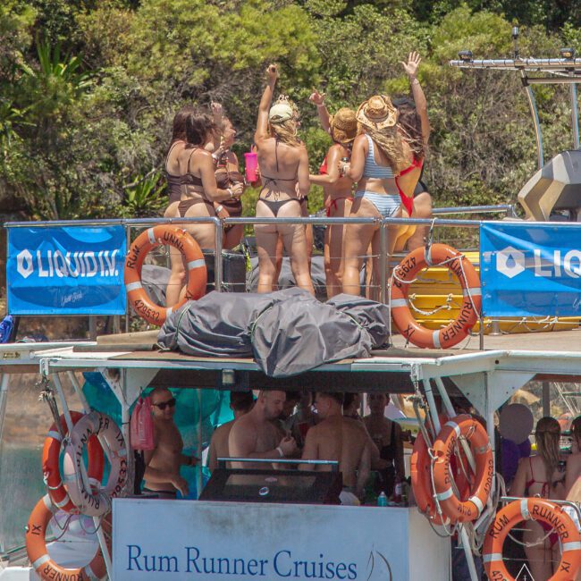 A group of women in swimsuits stand and wave on the upper deck of a Rum Runner Cruises boat, while people below relax in the shade; the boat is on the water near lush greenery.