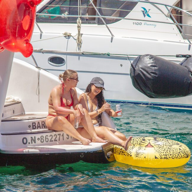 Two women in swimsuits relax on the back of a boat, dipping their feet in the water while sitting on the edge and a leopard-print float. A large white yacht and an inflatable toy are visible in the background.