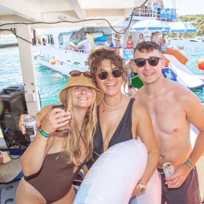 Three friends in swimsuits smile and pose together on a boat, holding drinks and a pool float. Other people and another boat with inflatable toys are visible on the water in the background. It's a sunny day.