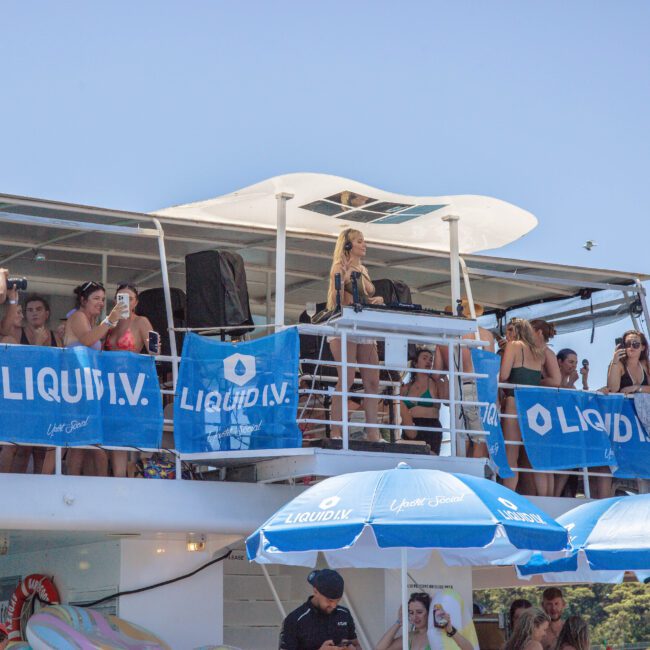 A group of people enjoy a party on a boat decorated with blue "LIQUID I.V." banners, with umbrellas and drinks, under sunny skies. Some guests are on the upper deck, taking photos and socializing.