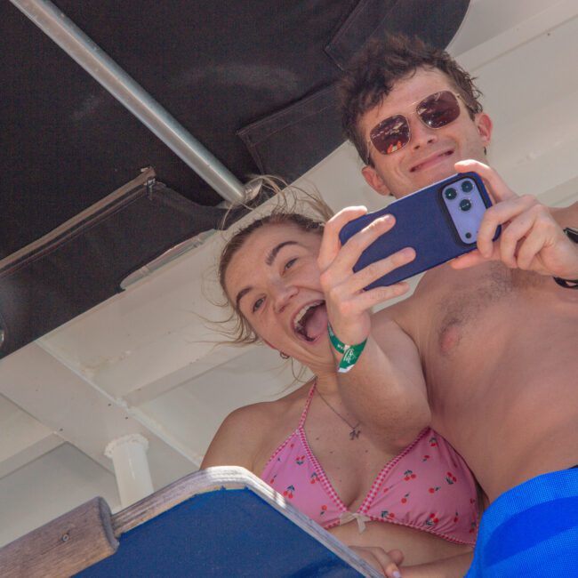 A smiling man and woman in swimwear take a selfie together on a boat, enjoying a sunny day. Another person in sunglasses is partially visible beside them. The Yacht Social Club logo is in the corner.