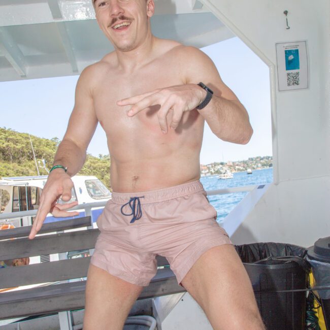 A young man in light pink swim trunks and a watch poses playfully on a boat, smiling and pointing with both hands. Blue water and coastal scenery are visible through the windows behind him.