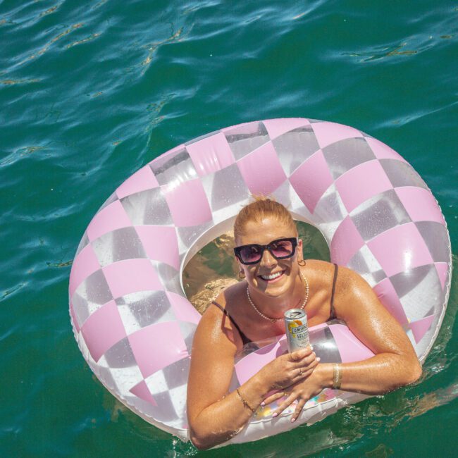 A woman with sunglasses smiles while relaxing on a pink and silver checkered inflatable ring in the water, holding a canned drink. The scene is sunny and bright. The logo "Yacht Social Club" appears in the bottom right corner.