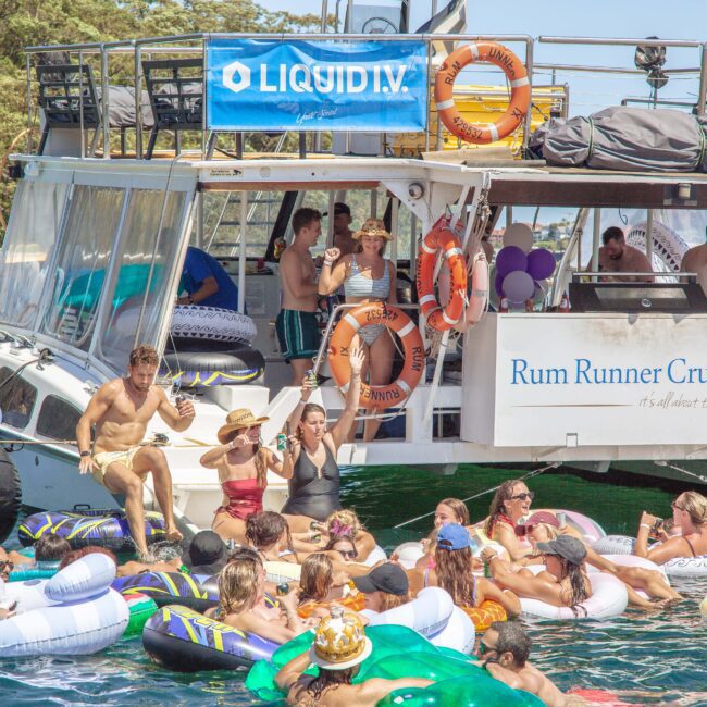 A group of people in swimsuits relax on colorful inflatables near a large, anchored party boat labeled "Rum Runner Cruises" on sunny water, with others socializing on the boat deck.