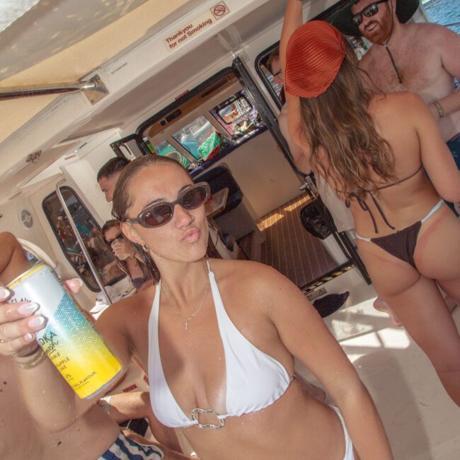 A woman in a white bikini and sunglasses poses with a drink can on a boat with several people in swimwear, enjoying a sunny day. Some people are dancing and talking; the yacht logo is visible in the corner.