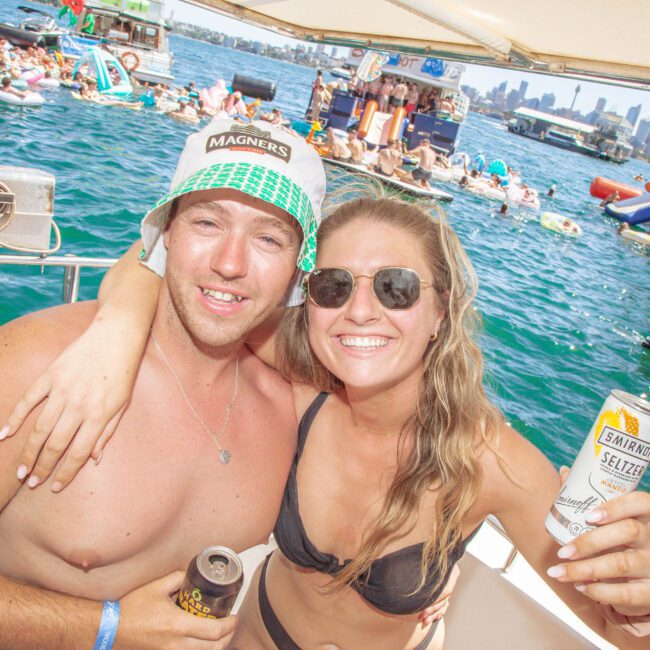 A man and woman in swimsuits smile and pose with drinks on a boat. The woman wears sunglasses, and both look happy. Behind them, a lively group of people float on inflatables in the water with city buildings in the distance.
