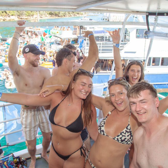 A group of young adults in swimwear smile and pose with their arms raised on a boat, with a crowd of people swimming and more boats visible on the bright, sunny water behind them.