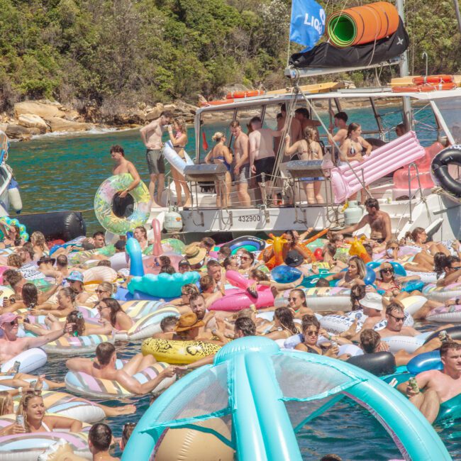 A large crowd of people on colorful inflatables gather in the water near boats, enjoying a sunny day at a lively floating party surrounded by trees and blue water.