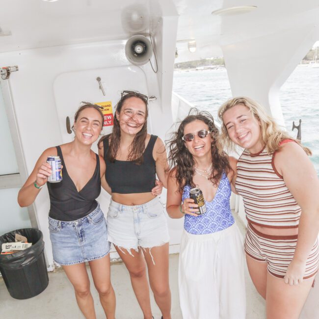 Four women smiling and posing together on a boat, with drinks in hand. The ocean and a coastline are visible in the background. They are dressed in summer clothes, enjoying a bright, sunny day.