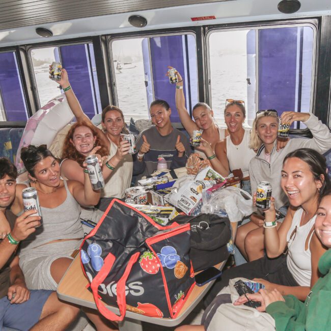 A group of smiling people sit around a table on a boat, raising canned drinks and snacks. Bright daylight streams through the windows, and the group appears cheerful and relaxed, enjoying their time together.