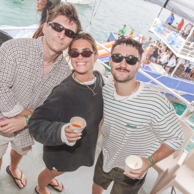 Three people in casual beachwear and sunglasses smile at the camera while holding drinks on a boat. The ocean and another boat with people are visible in the background. It appears to be a lively, sunny day.