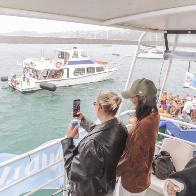 People on a boat party; a woman in sunglasses takes a photo with her phone, another person in a cap looks on, and several boats with groups of people are visible on the water in the background.