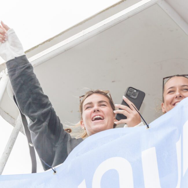 Two young women stand behind a blue and white banner on an elevated platform, smiling and cheering. One woman raises her arm, pointing upward, while both hold phones. The sky is bright and overcast.