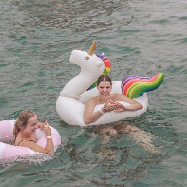 Two smiling women float in the water on inflatable pool toys—one shaped like a unicorn and the other a pink and white ring. Both are holding food and enjoying themselves. A wooden platform is visible on the right.