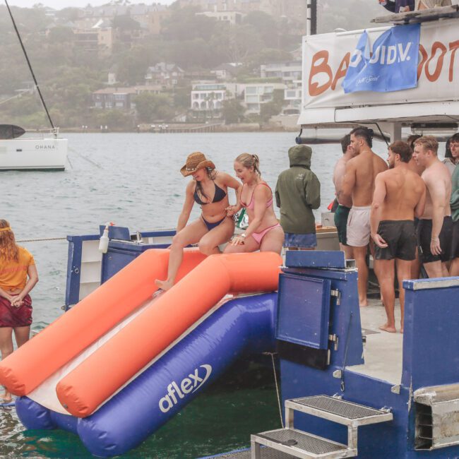 A group of people in swimwear gather on a boat with two orange inflatable slides. One person slides down while others wait. The boat is anchored on a lake, with houses and trees visible in the background.