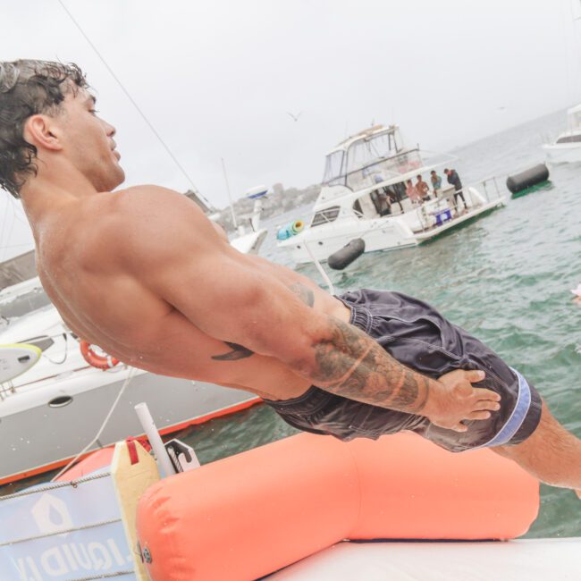 A muscular man in swim trunks leans back to jump or fall off a boat into the water, with yachts and people swimming in the background on a cloudy day.