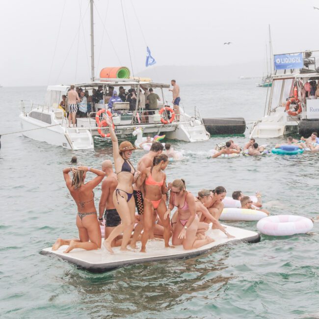 A group of people in swimsuits stand on a floating platform in the water near boats, while others relax on inflatable pool floats. The scene appears lively and social on a cloudy day.