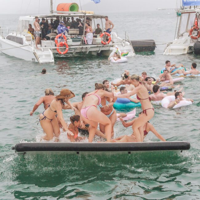 A group of people in swimsuits playfully pile onto a floating mat in the water near anchored boats, with others relaxing on inflatables. The scene is lively and festive, suggesting a fun summer gathering.