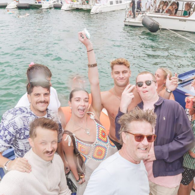 A group of people smiling and posing on a boat during a lively party, with others swimming and socializing in the water and on nearby boats under a cloudy sky. Everyone looks happy and relaxed.