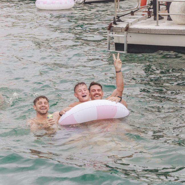 Three people smiling and posing in the water by a boat, one holding a striped inflatable ring and giving a peace sign, while another person swims nearby.