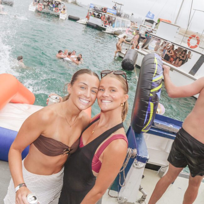 Two women smile at the camera on a boat during a lively party. Other people are swimming, floating on inflatables, and enjoying the water and nearby boats in the background. The vibe is festive and summery.