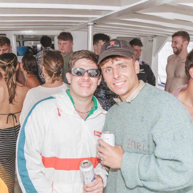 A group of young adults hang out on a crowded boat. Two men in the foreground smile at the camera holding drinks; one wears sunglasses and a jacket, the other a green sweater. People in swimwear mingle behind them.