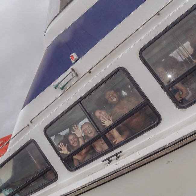 A group of people press their faces and hands against the windows of a white boat, smiling and laughing. The sky is overcast, and the boat has blue and orange accents. A "Yacht Social Club" logo appears in the corner.