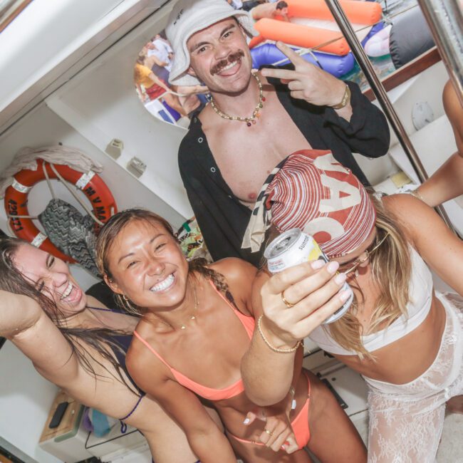 A group of five smiling people in swimsuits and summer outfits pose and celebrate on a boat, one person holds a drink and another flashes a peace sign; the scene is lively and joyful.
