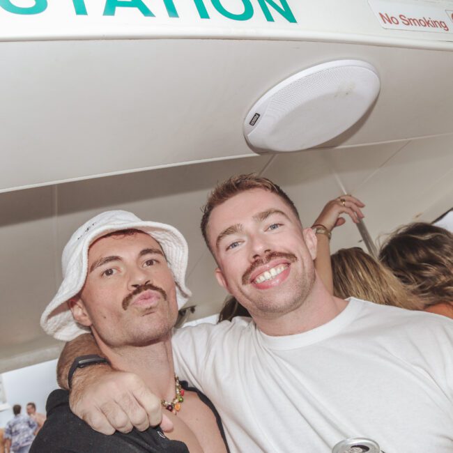 Two people smiling and posing together on a crowded boat. One wears a white bucket hat and necklace, the other a white T-shirt and holds a drink can. There are more people and party decorations in the background.
