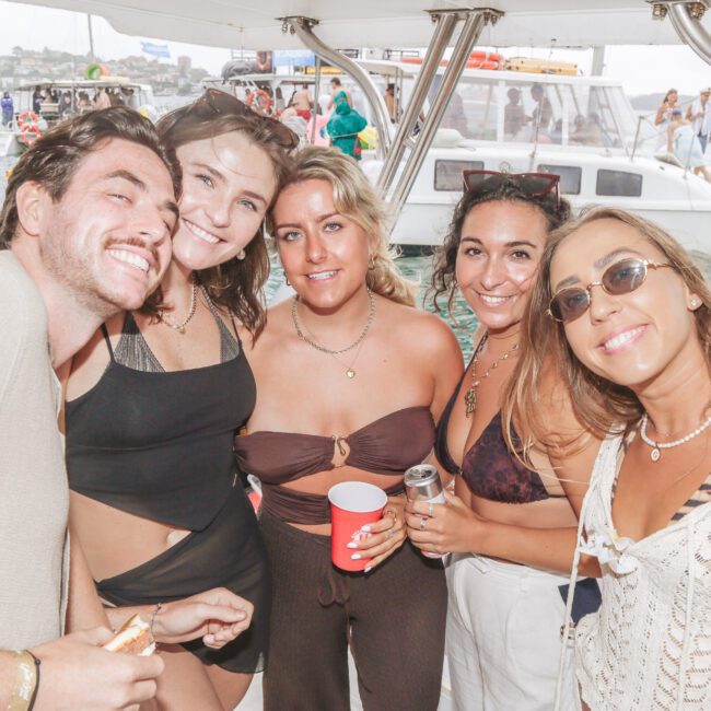 Five smiling young adults pose together on a boat, holding drinks, with other boats and people on the water in the background. The atmosphere is relaxed and festive.