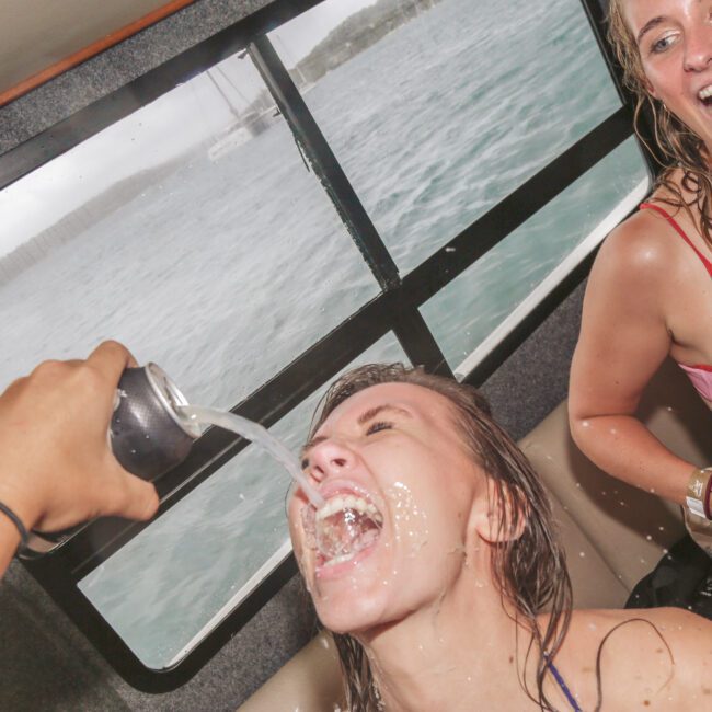 Two smiling women in swimsuits on a boat; one woman is being playfully sprayed in the mouth with a drink from a can while the other laughs. The sea is visible through the window behind them.