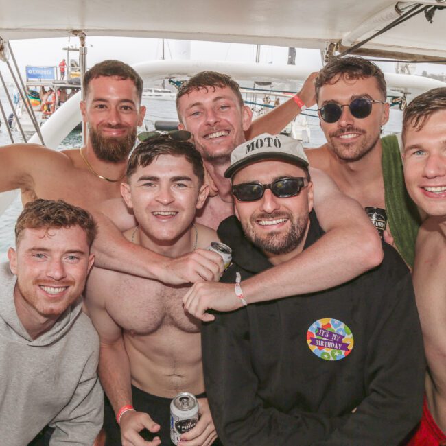 Seven young men are smiling and posing together on a boat, some shirtless, holding drinks. In the background, there is water, boats, and a cloudy sky. The atmosphere appears festive and relaxed.
