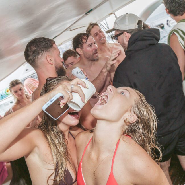 A group of young adults in swimsuits party on a boat. In the foreground, a woman pours a drink into another woman's mouth while others laugh and socialize in the crowded space.