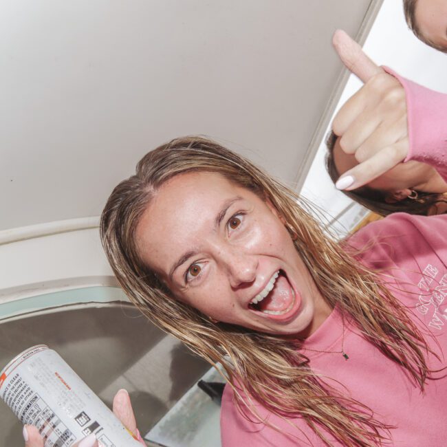 A young woman with wet hair, wearing a pink sweatshirt, smiles and makes a shaka hand sign. She holds a can, and two other people are partially visible in the background inside a boat. The "Yacht Social Club" logo is in the corner.