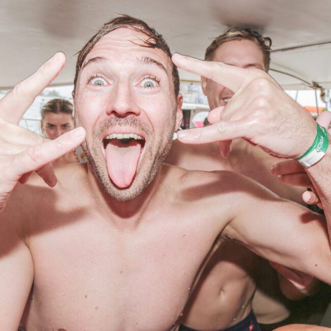 A shirtless man on a boat sticks out his tongue and makes rock-and-roll hand signs, smiling excitedly at the camera. Several other people, also in swimwear, are behind him, enjoying the festive atmosphere.