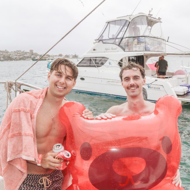 Two young men in swimwear smile on a boat, one holding a red inflatable bear pool float. A yacht and a shoreline with houses are visible in the background under an overcast sky.